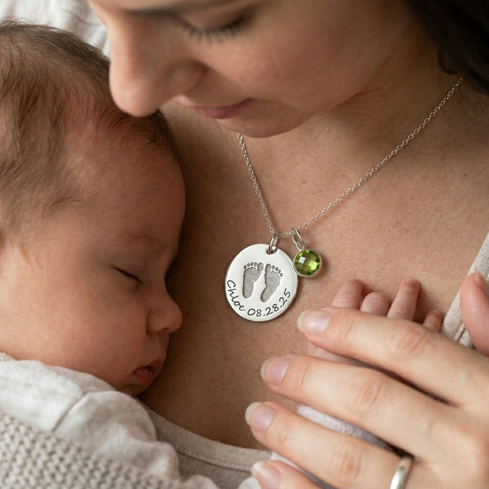 Mother wearing personalized baby footprint necklace with birthstone charm while holding a sleeping newborn.