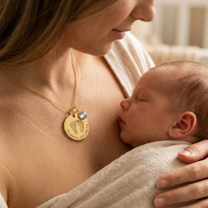 Mother holding newborn while wearing personalized baby footprint necklace with engraved name, date, and birthstone charm.