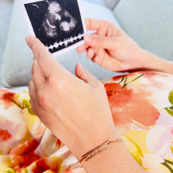 A person in a floral dress is wearing a personalized engraved heartbeat bracelet and looking at an ultrasound image with a sonogram. Each bracelet is crafted by hand and engraved with your actual image of your babys sonogram or ekg image.