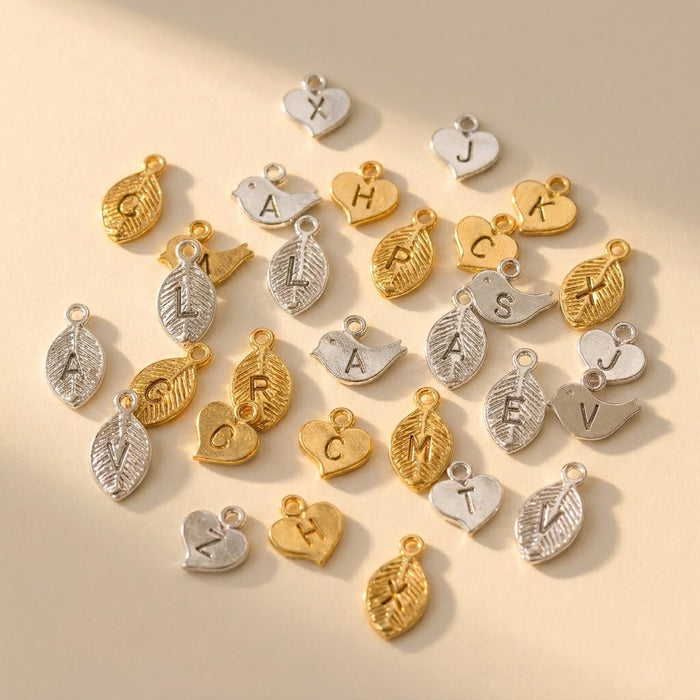 Hand stamped initial charms in gold and silver with leaf, bird, and heart shapes for adding a personalized charm to a necklace or bracelet.