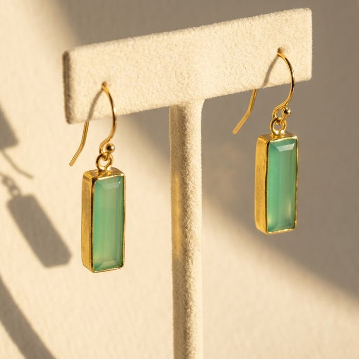Aqua chalcedony baguette bar earrings in gold displayed on a cream jewelry stand in natural sunlight.