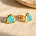 Aqua chalcedony triangle stud earrings in a gold bezel setting on cream linen in warm sunlight.