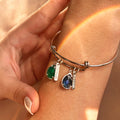 Close-up of a personalized silver family birthstone bangle with engraved name charms and green and blue teardrop gemstones.