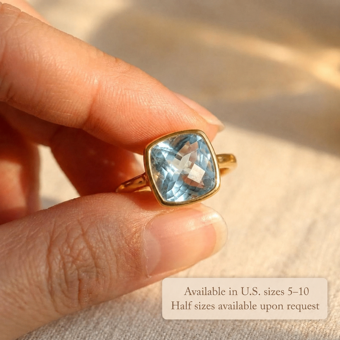 Close-up of cushion cut blue topaz ring held between fingers with text overlay showing U.S. ring sizes 5–10 and half sizes available upon request, perfect for customers choosing a December birthstone ring, blue gemstone ring, or custom size ring gift.