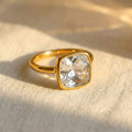 Cushion cut clear quartz ring in polished yellow gold bezel setting resting on soft neutral linen fabric, featuring a 9mm square transparent gemstone with diamond-like brilliance, April birthstone ring for women, minimalist modern fine jewelry, gold bezel set quartz ring, elegant stacking gemstone ring, handcrafted artisan birthstone jewelry, square clear gemstone ring gift for her
