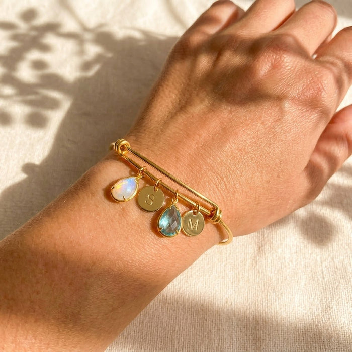 Custom family birthstone bangle bracelet on wrist with teardrop stones and initial charms in gold.