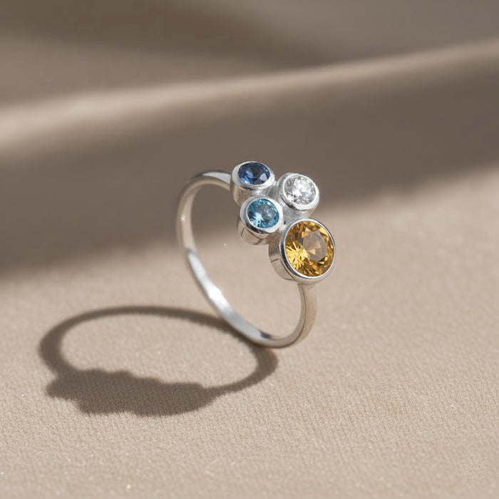 three stone birthstone ring resting on champagne satin fabric with warm light highlighting the gold vermeil finish and vibrant gemstones, personalized mom ring with three birthstones representing each child, elegant cluster style ideal for everyday wear or special occasions, thoughtful Mother’s Day gift, birthday gift or family jewelry heirloom crafted in sterling silver or gold vermeil with customizable stones