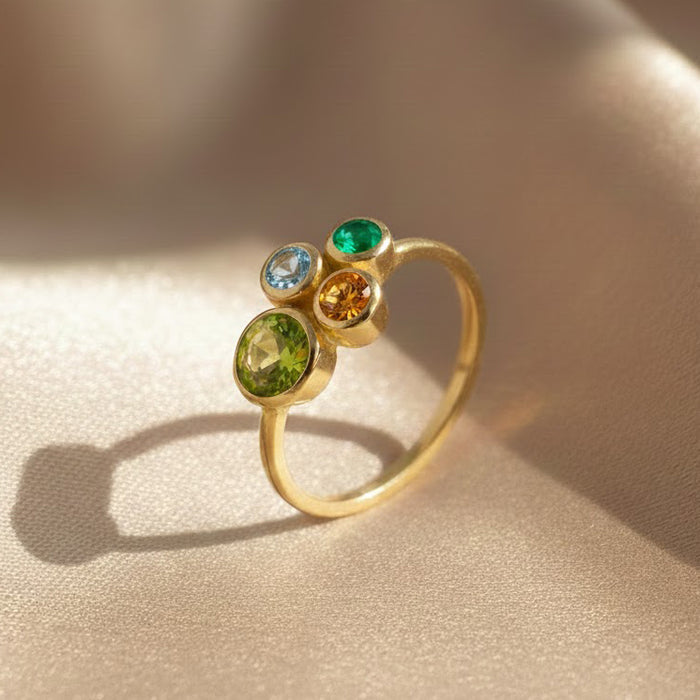 custom birthstone ring in matte gold vermeil featuring four bezel set gemstones in bright green, light blue, golden yellow and rich emerald tones, clustered together on a slim band and casting a soft shadow on champagne satin, elegant family birthstone ring that can be customized with children’s stones for mom or grandma, dainty yet colorful mothers ring perfect for everyday wear and for gifting on Mother’s Day, birthdays, anniversaries or Christmas