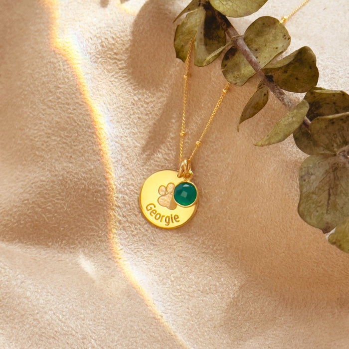 Engraved paw print disc necklace with emerald green birthstone charm on gold chain, styled on satin fabric background. Personalized jewelry gift for pet lovers, moms, and grandmas. Ideal Christmas gift, pet memorial keepsake, or everyday necklace. Features sterling silver, gold-filled, and rose gold-filled finishes with 12 birthstone options. Elegant dainty jewelry for birthdays, anniversaries, or holiday gifting.