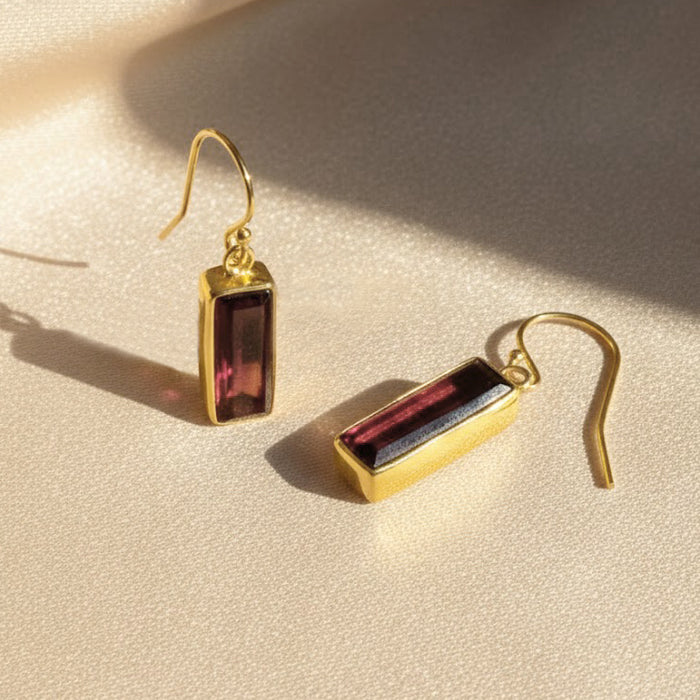 Garnet baguette drop earrings displayed on soft neutral fabric, featuring deep red rectangular garnet gemstones set in gold. Minimalist birthstone earrings designed with a slim baguette cut for elegant everyday wear. Timeless garnet earrings perfect as January birthstone jewelry, anniversary gift, birthday present, or meaningful gemstone earrings for women.