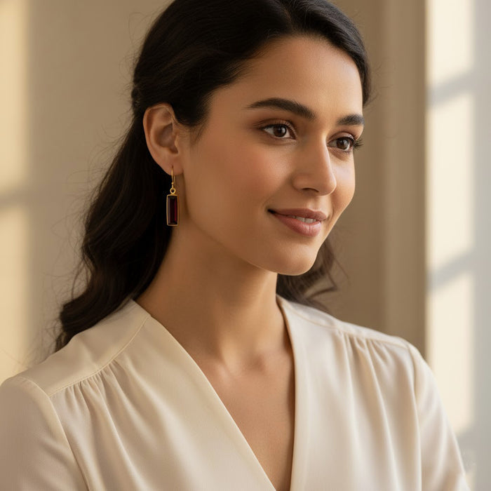 Woman wearing garnet baguette earrings indoors, showcasing delicate rectangular garnet drop earrings in gold. Elegant minimalist gemstone earrings designed for everyday wear and special occasions. January birthstone earrings that make a thoughtful gift for women who love refined, understated jewelry with a modern silhouette.