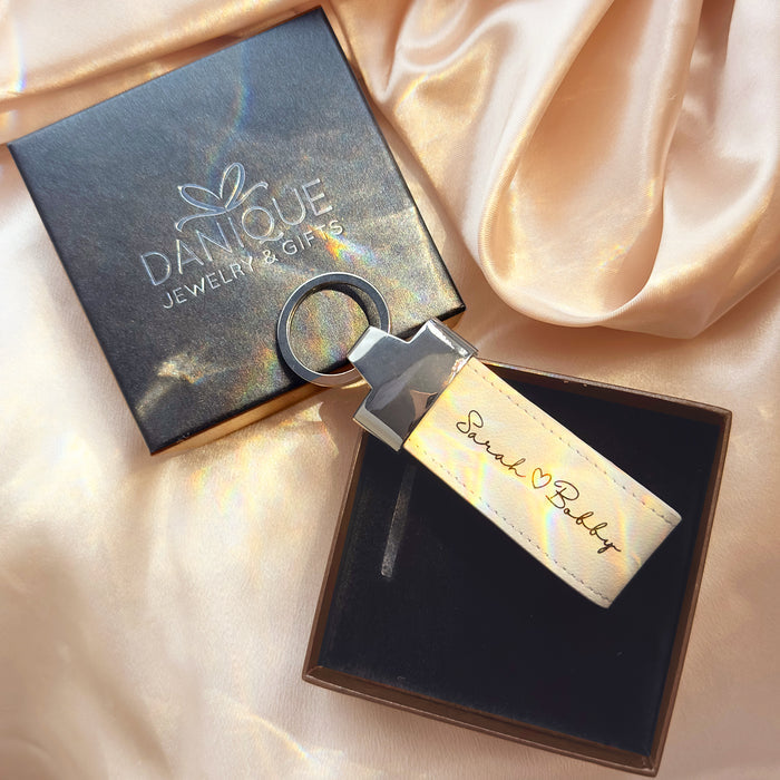 Personalized large leather keychain loop in ivory cream color engraved with “Sarah ♥ Bobby,” displayed in a black gift box beside a Danique Jewelry & Gifts branded box on soft satin fabric. This custom leather keychain makes a romantic gift for couples, perfect for anniversary, long distance relationship gift, or Valentine’s Day present. Ideal for boyfriend or girlfriend gifts with engraved names or coordinates. Durable stitched leather key ring accessory for everyday use or sentimental keepsake.