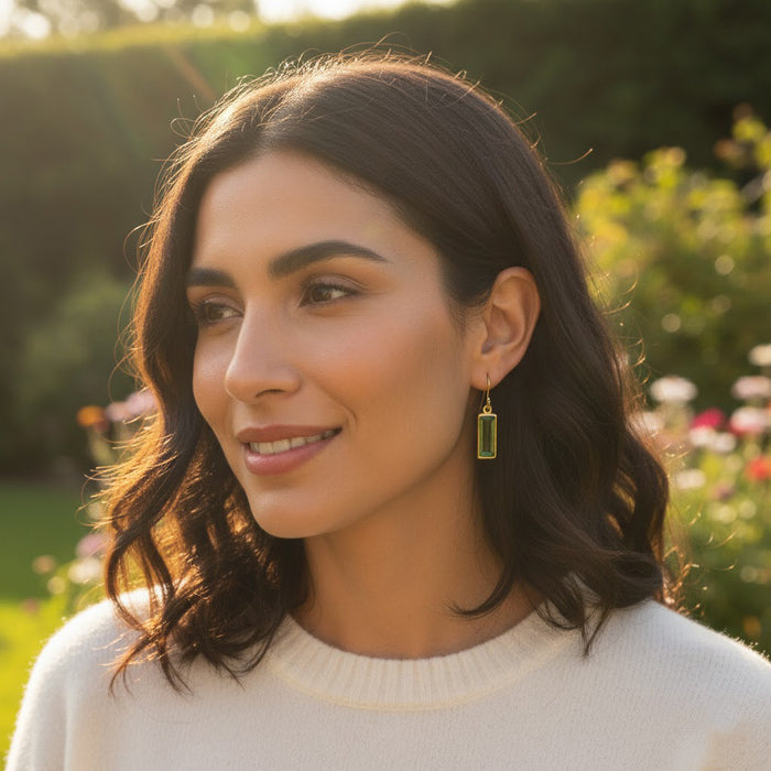 woman wearing a single baguette cut birthstone earring in rich green on a sunny garden afternoon, elongated rectangular gemstone bezel set in warm gold vermeil and hanging from a delicate French hook, modern minimalist statement earring that adds a subtle pop of color, perfect as personalized birthstone jewelry for birthdays, anniversaries or holiday gifts, designed to pair with matching baguette bar necklaces or to wear solo for elegant everyday style