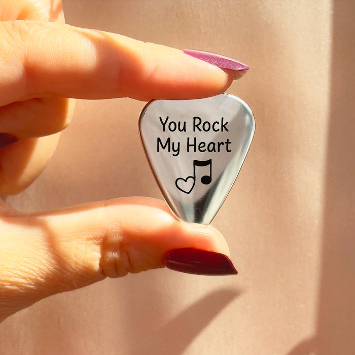 Close-up of hand holding a stainless steel guitar pick engraved with the phrase “You Rock My Heart” and a music note icon, showing the polished surface and comfortable triangular shape. Romantic custom guitar pick for boyfriend, husband or partner, an ideal Valentine’s Day, anniversary or birthday gift for musicians and music lovers, personalized plectrum that is both playable and a lasting keepsake.