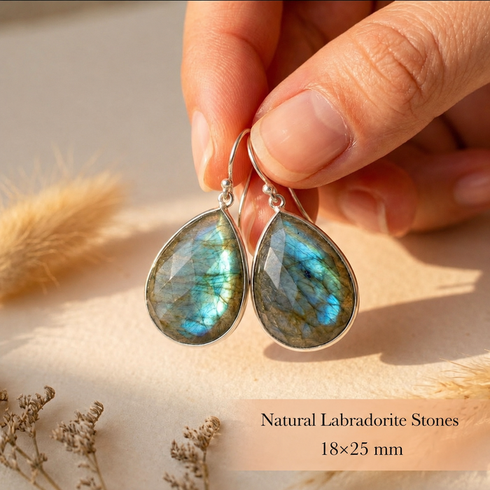 Hand holding large labradorite teardrop earrings, showing the natural flash, faceted stone surface, and gold ear wires for a close-up size reference.