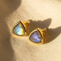 Labradorite triangle stud earrings in yellow vermeil gold with faceted blue green gemstone flash on warm neutral fabric.