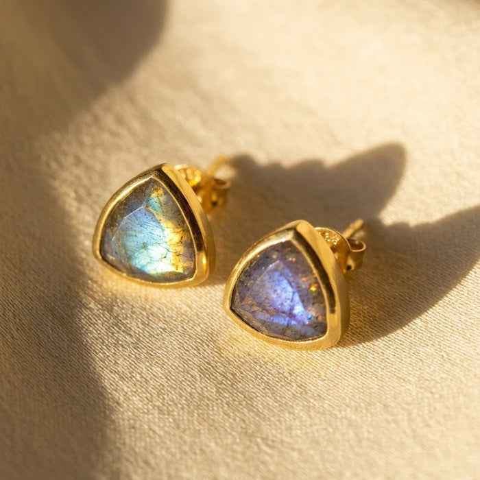 Labradorite triangle stud earrings in yellow vermeil gold with faceted blue green gemstone flash on warm neutral fabric.