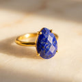 Natural lapis lazuli teardrop ring in gold prong setting with faceted deep blue gemstone on a warm neutral background.