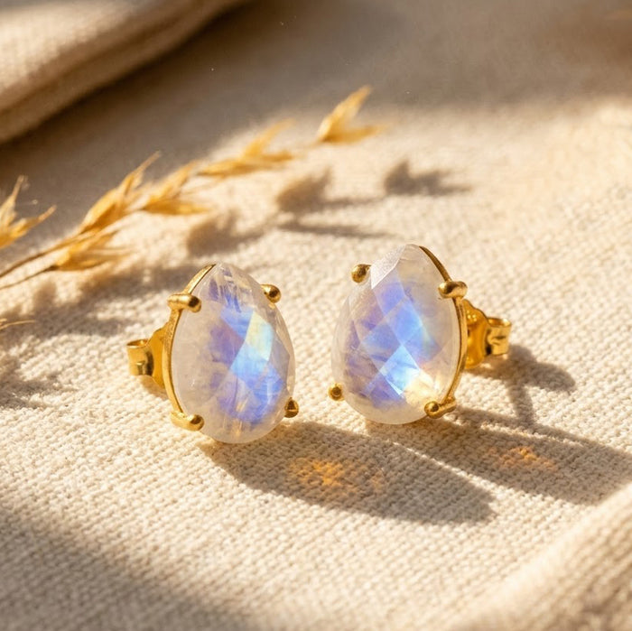 Teardrop rainbow moonstone stud earrings in gold with prong settings, displayed on warm cream fabric with natural blue and white moonstone flashes.