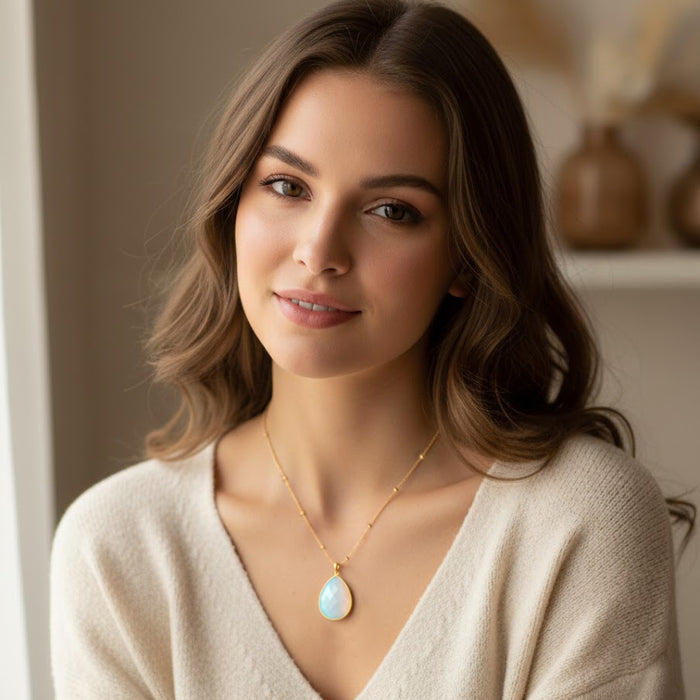 A dainty opalite necklace with a teardrop gemstone pendant on a gold chain worn by a woman in soft natural light. The minimalist opal pendant shines with subtle iridescence, perfect for bridesmaid gifts, bridal jewelry, everyday gemstone necklaces, and meaningful birthday gifts. Designed for effortless elegance, layering, and timeless modern style in opalite jewelry.
