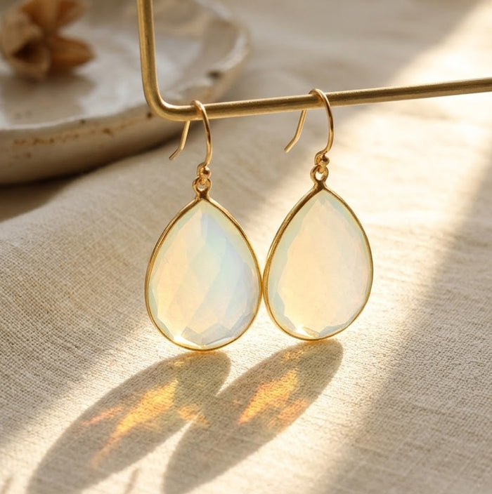 Close-up of opalite teardrop earrings in gold with soft iridescent flashes and warm shadows.