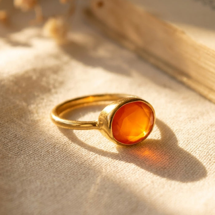 Natural oval carnelian ring in yellow gold vermeil displayed on cream linen in warm sunlight, showing the faceted orange gemstone and bezel setting.