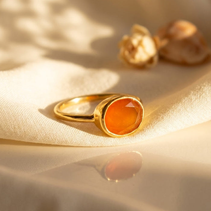 Close-up of a natural faceted oval carnelian ring in a polished yellow gold vermeil bezel setting on softly folded neutral fabric.