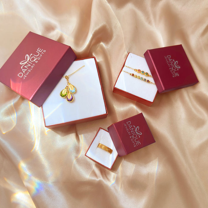 Danique Jewelry ready-to-gift packaging on champagne satin showing a gold teardrop birthstone pendant necklace, two dainty layered gemstone bracelets, and a textured gold ring nestled in red logo boxes with white inserts. Elegant handmade pieces with customizable stones and metals, ideal as a meaningful gift for her. Perfect for birthdays, anniversaries, Mother’s Day or Christmas, highlighting premium presentation, keepsake quality, and easy unboxing for shoppers seeking personalized jewelry.