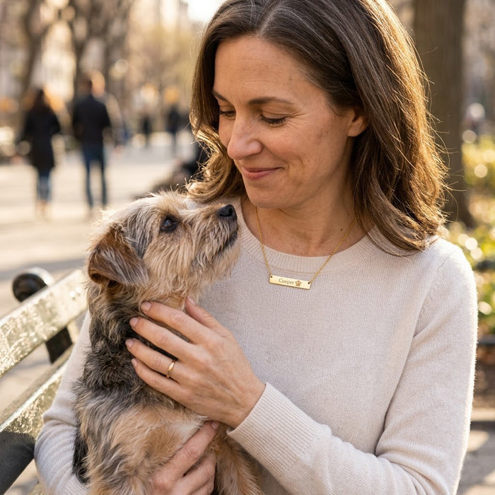 Woman wearing personalized paw print bar necklace while holding a small dog, custom pet memorial jewelry for dog lovers.