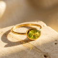 Dainty oval peridot ring in a gold bezel setting, shown in warm sunlight on a natural stone surface.