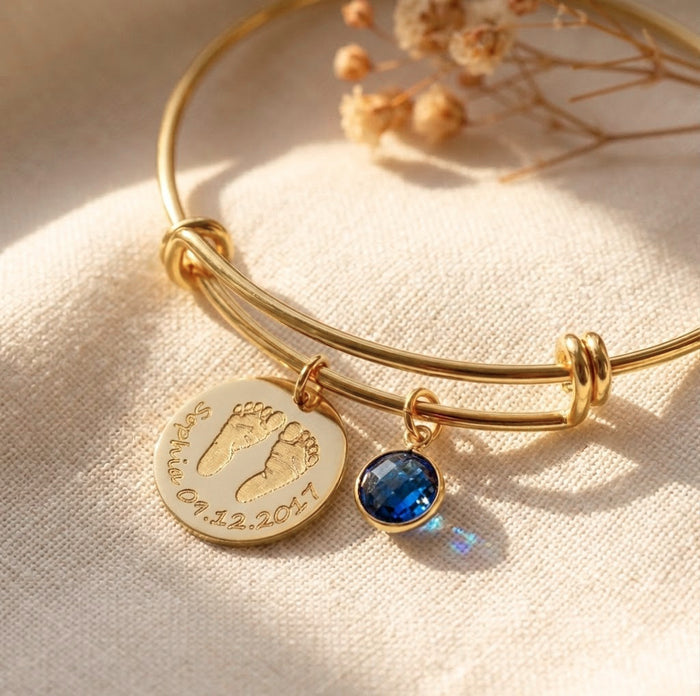 Close-up of custom baby footprint bangle with engraved name, date, and blue birthstone charm on gold adjustable bracelet.