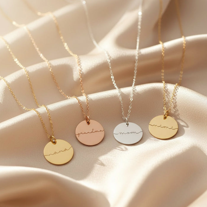 Personalized name disc pendant necklaces in gold, rose gold and sterling silver shown on soft satin background, engraved with “nana”, “grandma”, “mom” and “mimi”; custom delicate jewelry gift for mothers, grandmothers, Mother’s Day or birthdays