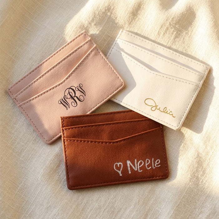 Personalized PU leather card holders in pink, white, and brown with engraved names and monograms on a warm neutral background.