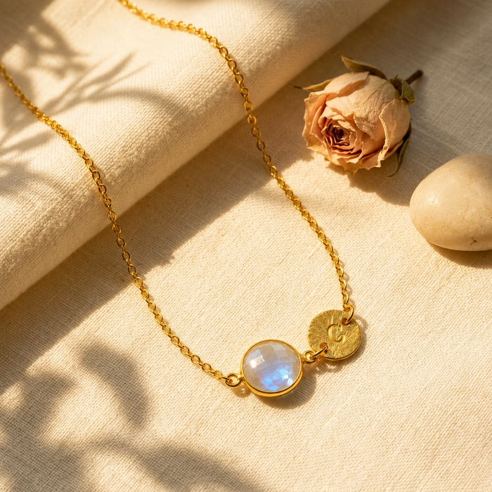 Gold personalized moonstone initial necklace with round faceted June birthstone displayed on cream fabric.