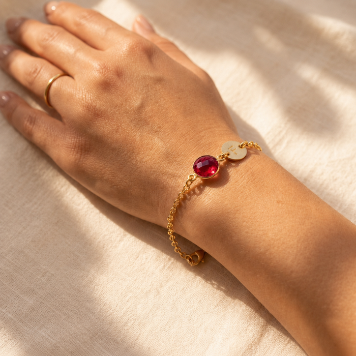 Gold ruby initial bracelet worn on a wrist in warm natural sunlight