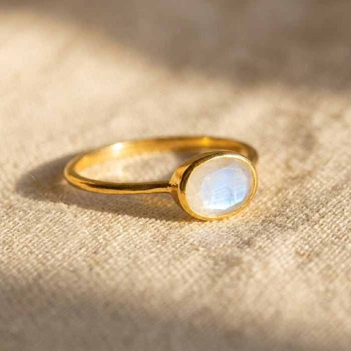 Small oval rainbow moonstone ring in yellow gold with subtle blue flash, shown close up on warm neutral fabric.