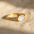 Small oval rainbow moonstone ring in yellow gold with subtle blue flash, shown close up on warm neutral fabric.