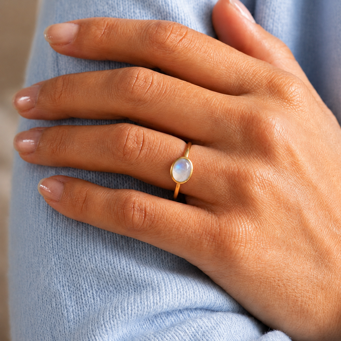 Oval rainbow moonstone ring worn on a model’s hand with a soft blue sweater background.