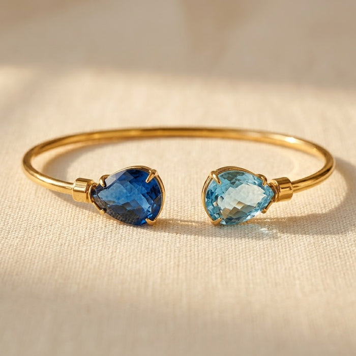 Adjustable two birthstone bangle bracelet with blue sapphire and light blue topaz teardrop gemstones on a warm neutral fabric background.