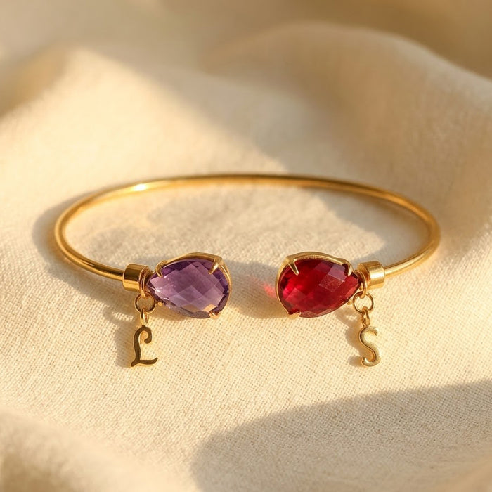 Two birthstone initial bangle bracelet in gold with purple and red teardrop gemstones and matching letter charms, personalized for two loved ones.