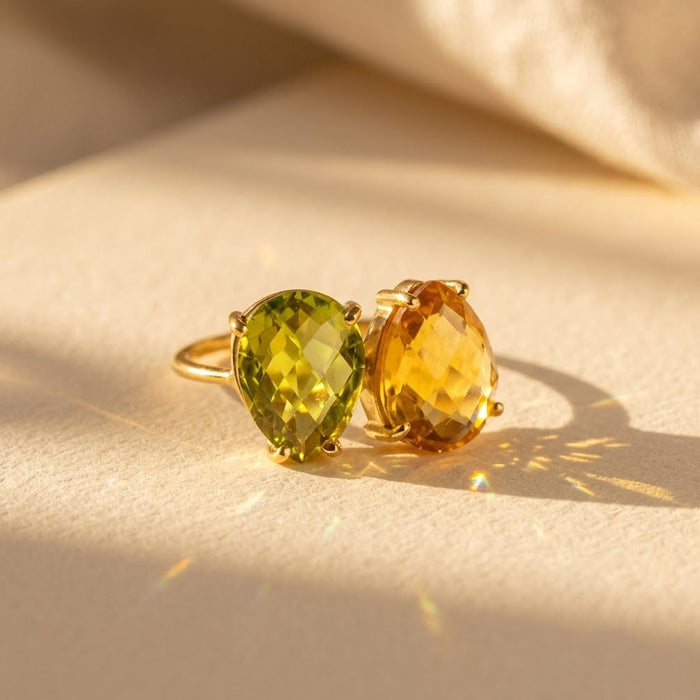 Two birthstone teardrop ring with green and yellow faceted stones on a warm neutral background