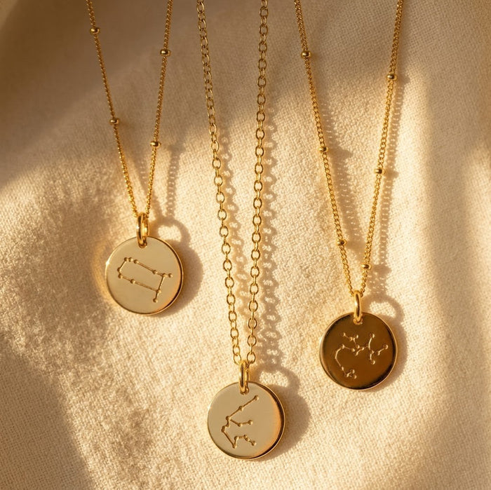 Zodiac constellation necklaces in sterling silver, yellow gold, and rose gold finishes on a neutral fabric background.
